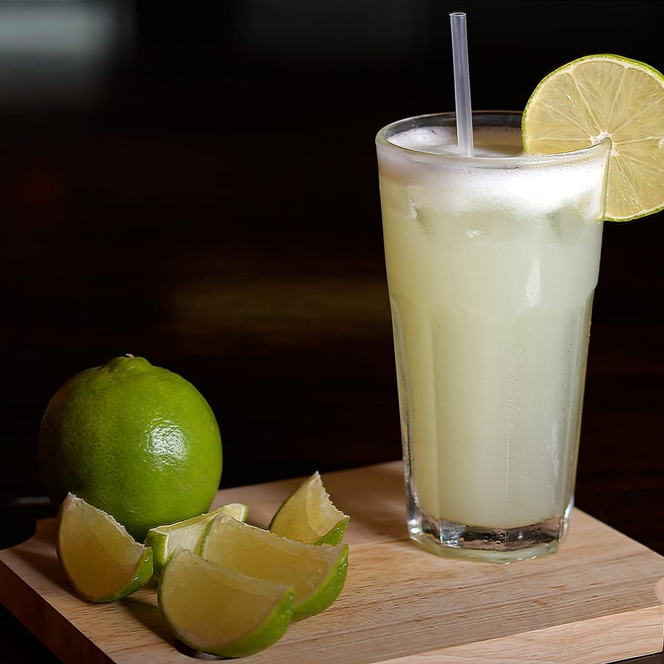Tall glass of pale lime drink with straw and lime slice, surrounded by whole and wedged limes on a wooden board.