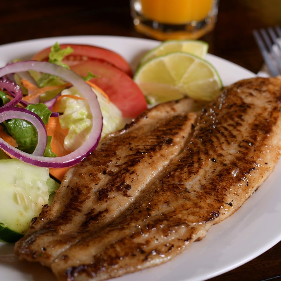 Grilled seasoned fish fillet served with fresh salad, cucumber, tomato, lime wedges, and an orange drink.