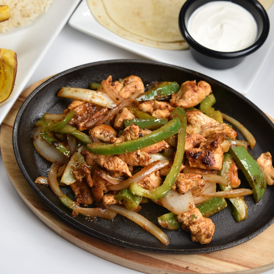 Sizzling chicken fajitas with green peppers and onions served with tortillas, sour cream, rice, and fried plantains.