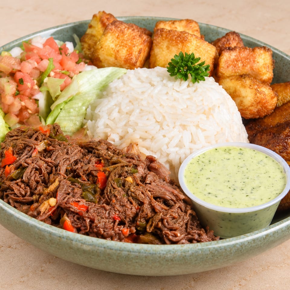 Bowl of white rice with shredded beef, fried plantains, crispy cheese, salad, and green sauce.