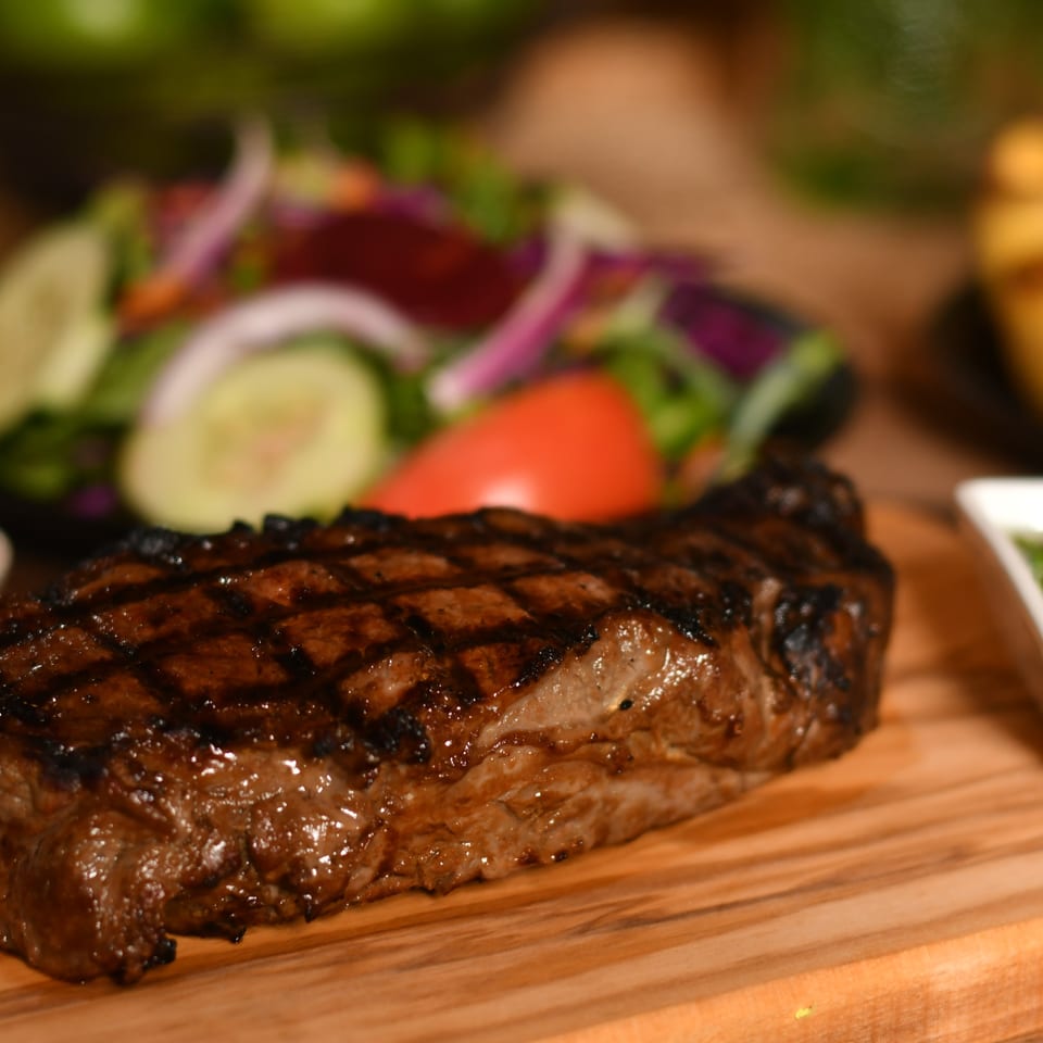 Grilled steak with charred grill marks served on a wooden board alongside salad, sauces, and vegetables.